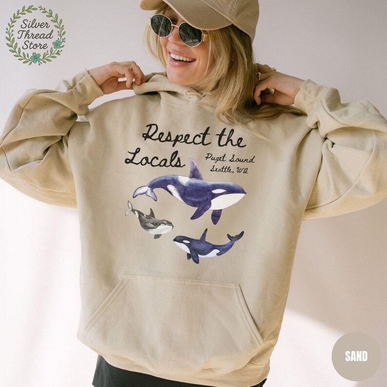 May include: Sand-colored hoodie featuring a graphic of orcas with the text "Respect the Locals" and "Puget Sound, Seattle, WA." The design includes three orcas in shades of blue, black, and white. The hoodie has a front pocket and a drawstring hood.