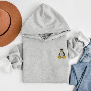 May include: A light gray hoodie with a hood and long sleeves. The hoodie features a small embroidered penguin on the front. The sleeves are cuffed with white fabric. A brown hat and blue jeans are also in the image.