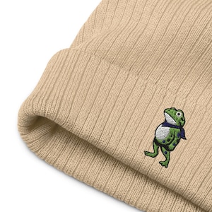 May include: A beige ribbed knit beanie featuring an embroidered green frog wearing a blue scarf. The frog has white eyes and a white belly with green spots. The beanie is made of a soft, warm material.