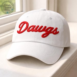 May include: White baseball cap with the red embroidered word "Dawgs" across the front. The cap has a curved brim and a button on top. The hat is sitting on a white surface.