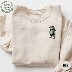 May include: Sand-colored sweatshirt with an embroidered frog design. The frog is green with black spots and a blue scarf. The sweatshirt has a crew neck and the text "Silver Thread Store" is visible in the upper left corner.