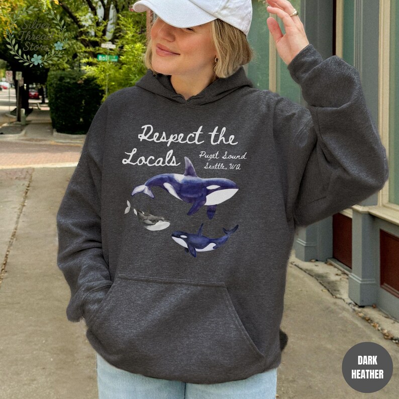 May include: Dark heather gray hoodie with a front pocket. The hoodie features a graphic of orca whales and the text "Respect the Locals Puget Sound Seattle, WA". The hoodie is made of a soft material.