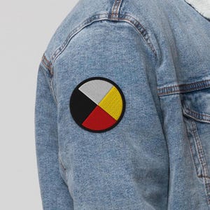 May include: A blue denim jacket with a circular patch on the left sleeve. The patch is made of four coloured sections: black, red, yellow, and white. The colours are arranged in a circle, with the black section at the top.