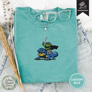 Peut inclure: T-shirt bleu lagon avec un motif brodé d'une grenouille guerrière chevauchant un dragon bleu, tenant une épée. Le t-shirt a un col rond et des manches courtes. Un collier argenté et des lunettes transparentes sont posés sur le t-shirt.