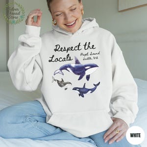 May include: A white hoodie with a graphic of three orcas and the text "Respect the Locals" and "Puget Sound, Seattle, WA". The orcas are in shades of gray and blue. The hoodie has a front pocket and a drawstring hood.
