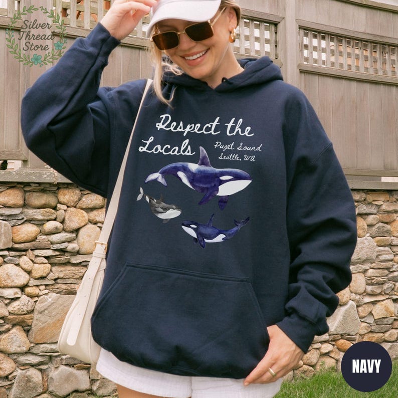 May include: Navy blue hoodie with a graphic of orcas and the text "Respect the Locals Puget Sound, Seattle, WA." The hoodie has a front pocket and a drawstring hood. The word "NAVY" is printed on the bottom right.