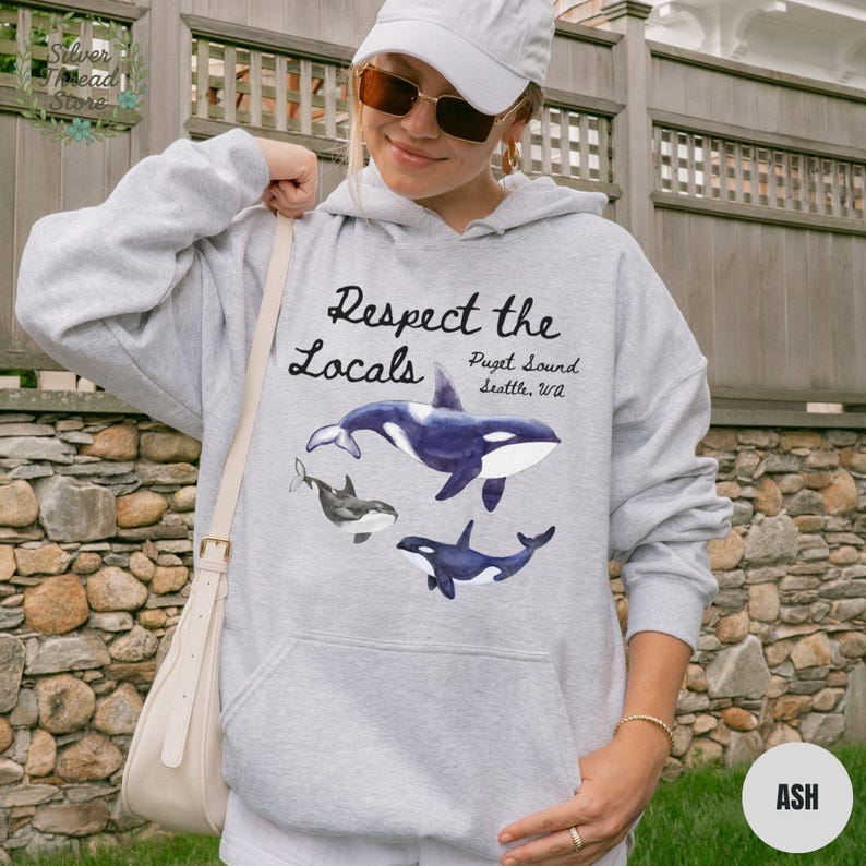 May include: Light gray hoodie with a graphic of three orcas and the text "Respect the Locals Puget Sound Seattle, WA." The hoodie has a drawstring hood and a front pocket. The word "ASH" is printed on a small, round tag.