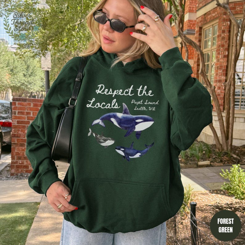 May include: Forest green hoodie with the text "Respect the Locals" and illustrations of orcas. The hoodie also includes the text "Puget Sound, Seattle, WA". The hoodie is a solid forest green color.