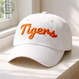May include: A white baseball cap with the word "Tigers" embroidered in orange script across the front. The cap has a curved brim and a button on top. The cap is sitting on a white surface.