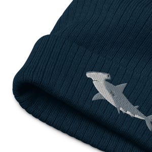 May include: A dark blue knitted beanie with a white embroidered hammerhead shark design.