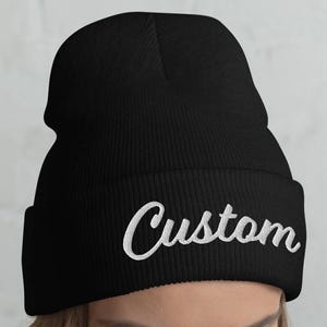May include: Black beanie with the word "Custom" embroidered in white script across the front. The beanie is made of a soft, knitted material and is designed to be worn on the head.