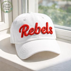 May include: White baseball cap with the red word "Rebels" embroidered on the front. The cap has a curved brim and a button on top. The hat is sitting on a white surface near a window.