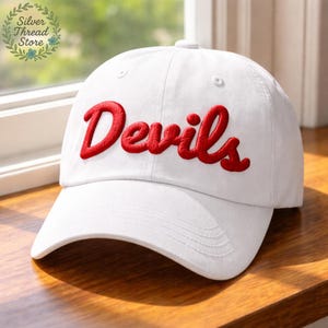 May include: White baseball cap with the red word "Devils" embroidered on the front. The cap has a curved brim and a button on top. The hat is made of a soft material and is suitable for casual wear.