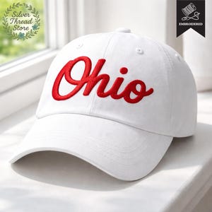 May include: A white baseball cap with the word "Ohio" embroidered in red script across the front. The cap has a curved brim and a button on top. The cap is made of a soft, durable material.
