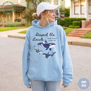 May include: Light blue hoodie with a graphic of orcas and the text "Respect the Locals Puget Sound Seattle, WA." The hoodie has a front pocket and a drawstring hood. The text "LIGHT BLUE" is in the bottom right corner.