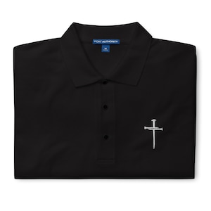 May include: Black polo shirt with a white embroidered cross with nails on a black background. The shirt has a blue label that says "PORT AUTHORITY" and "M".