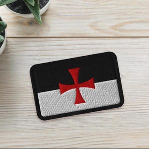 May include: Embroidered patch featuring a red cross on a black and white background. The patch has a rectangular shape with a black border. The design is likely a symbol or emblem.