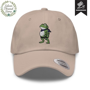 May include: A tan baseball cap with an embroidered frog design. The frog is green with blue spots and a blue scarf. The cap has an adjustable strap and a curved brim. The cap is from the Silver Thread Store.
