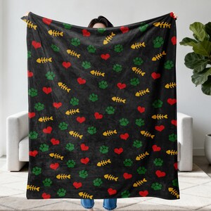 May include: Black blanket with a pattern of red hearts, green paw prints, and yellow fish bones. The blanket is held up, showcasing the repeating design. The blanket is a cozy item for cat lovers.