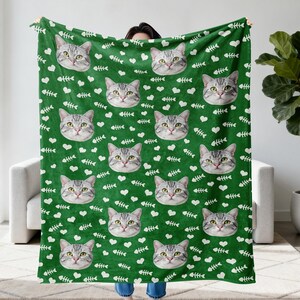 May include: A green blanket featuring a repeating pattern of cat faces, white hearts, and fish bones. The cat faces are gray and white, with yellow eyes. The blanket is made of a soft material and is perfect for cat lovers.