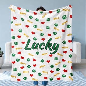 May include: White blanket with a pattern of green paw prints, red hearts, and yellow fish bones. The word "Lucky" is written in green script. The blanket is held up in front of a person.