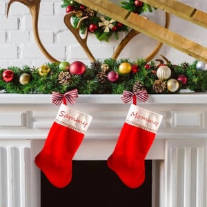 May include: Two red Christmas stockings with white tops hang from a white mantel. The stocking on the left says "Sammy" and the one on the right says "Mommy". A garland of greenery, ornaments, and pinecones decorates the mantel.