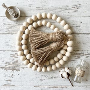 May include: A decorative wooden bead garland with two jute tassels. A small glass bottle with "ARTISAN DECOR" text, a cotton stem, and a small ceramic bowl with a bundle of sage are also present. The items are arranged on a white, distressed wooden surface.