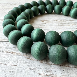 May include: Close-up of a strand of dark green wooden beads. The beads are round and have a matte finish. The beads are strung together, forming a curved shape. The background is a light-colored, textured surface.