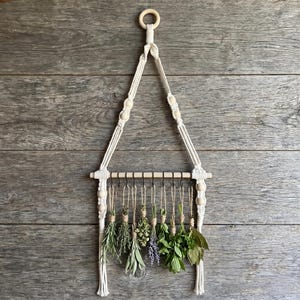 May include: A macrame herb drying rack with a wooden dowel and hanging loops. The rack is adorned with various dried herbs, including rosemary, thyme, and basil. The macrame is cream-colored, and the herbs are green and purple.