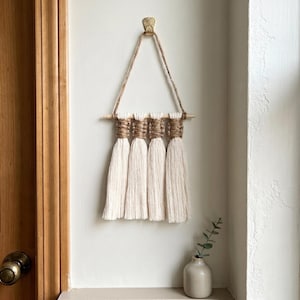 May include: A handmade macrame wall hanging featuring four off-white tassels suspended from a natural wooden dowel. The top section is wrapped with tan-colored twine, and the piece hangs from a jute cord. The wall decor is mounted on a white wall.