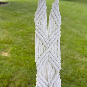 May include: A white macrame plant hanger featuring a series of intricate, hand-knotted geometric diamond patterns. The cord is thick and textured, forming a sturdy loop at the top for hanging, with long, straight fringe extending downward from the knots.