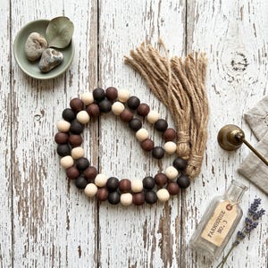 May include: A decorative wooden bead garland in shades of brown, black, and cream, with a jute tassel. The garland is arranged on a distressed white wooden surface, alongside a small dish with stones, a vintage bottle, and sprigs of lavender.