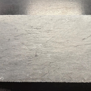May include: Rectangular gray slate serving board with a rough, textured surface. The board is a neutral color and is suitable for serving appetizers or small dishes. The edges are uneven, adding to its rustic appearance.