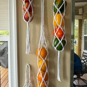 May include: Four white macrame hanging fruit hammocks suspended vertically. The hammocks contain various fresh produce including red apples, oranges, yellow and green bell peppers, and white garlic bulbs, each featuring knotted cord designs and tassels.