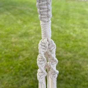May include: An off-white macrame plant hanger featuring a top loop attached to a black carabiner, a thick braided section, and multiple knotted cords extending downward with small wooden beads integrated into the lower hanging strands.