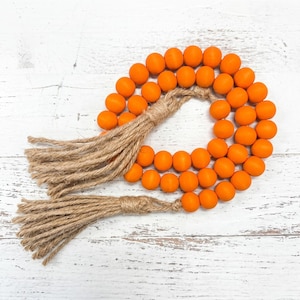 May include: A decorative garland featuring bright orange wooden beads. The beads are strung together and accented with natural jute tassels at each end. The garland is arranged in a circular shape, perfect for home decor.