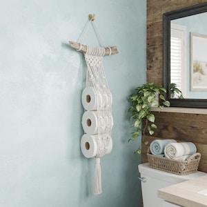 May include: A macrame toilet paper holder made of off-white cotton cord featuring a knotted net design suspended from a piece of driftwood. The holder contains three rolls of white toilet paper stacked vertically and finishes with a long tassel.