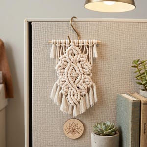 May include: A cream-colored macrame wall hanging with a detailed geometric design. It is suspended from a gold-colored hook and wooden dowel. The piece has multiple tassels at the bottom. A wooden coaster with a lotus flower design is visible.