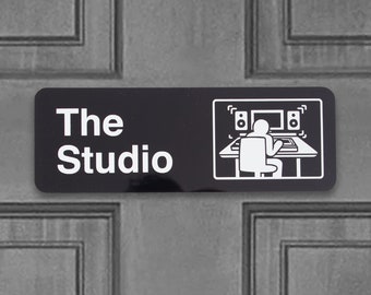 May include: A black and white sign with the text "The Studio" and a graphic of a person sitting at a desk with two computer monitors and speakers.