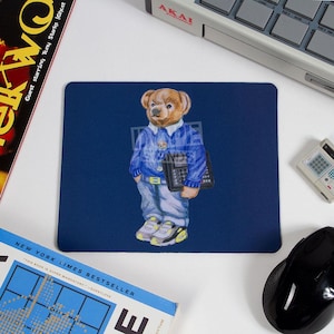 Peut inclure: Un tapis de souris rectangulaire bleu marine présente une illustration à l'aquarelle d'un ours en peluche portant un pull bleu, un jean et des baskets, tenant une calculatrice. Le tapis est entouré d'accessoires informatiques et d'un livre.