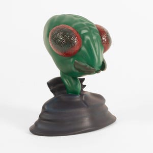 May include: A green and red figurine of a creature with large eyes and a textured surface. The creature is wearing a dark blue jacket and is standing on a dark blue base.