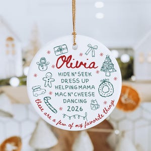 May include: White ceramic Christmas ornament with gold string. The ornament features the name "Olivia" in red script, surrounded by festive doodles and text: "Hide N' Seek, Dress Up, Helping Mama, Mac N' Cheese, Dancing, 2026".