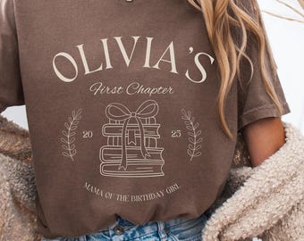 May include: A brown t-shirt with the text "Olivia's First Chapter" in white. The design features a stack of books with a bow on top and the numbers 20 and 25 on either side. The text "Mama of the Birthday Girl" is below the books.