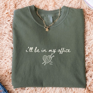 May include: Moss green t-shirt with the text "I'll be in my office" and a yarn and needle graphic. A gold chain necklace rests on the shirt. White sneakers and blue jeans are also visible.