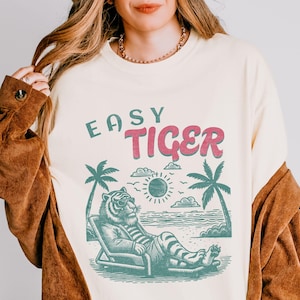 May include: A cream coloured t-shirt with the text "Easy Tiger" in pink and blue lettering. The graphic features a cartoon tiger wearing sunglasses and relaxing on a beach chair.