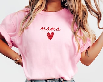 Könnte beinhalten: Ein hellrosa T-Shirt mit kurzen Ärmeln, das in der Mitte der Brust das Wort mama in einer roten Schreibschrift zeigt, direkt über einer kleinen, handgezeichneten roten Herzgrafik. Das Shirt hat einen Rundhalsausschnitt und kurze Ärmel.