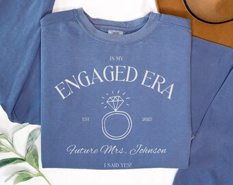 May include: A blue crewneck sweatshirt featuring white text that reads IN MY ENGAGED ERA, EST 2023, Future Mrs. Johnson, and I SAID YES!, along with a line-art graphic of a diamond ring centered on the chest of the garment.