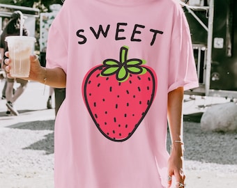 May include: A light pink short-sleeved t-shirt featuring the word SWEET printed in black capital letters above a large graphic of a red strawberry with a green stem and black seeds. The shirt has a crew neckline and a loose, relaxed fit.