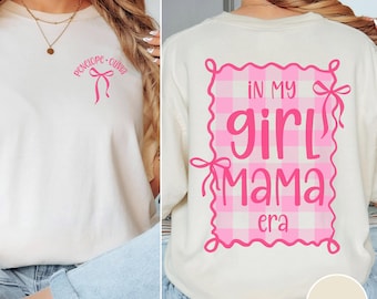 May include: An ivory t-shirt featuring a pink bow graphic and the text "penelope • olivia" on the front chest. The back displays a pink gingham rectangular frame with bows at the corners and the text "IN MY girl MAMA era" in pink lettering.