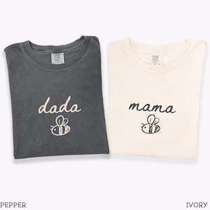 May include: Two folded t-shirts, one in pepper and one in ivory. The pepper shirt features the word "dada" and a bee illustration. The ivory shirt displays the word "mama" and a bee illustration. Both shirts are made of a soft fabric.